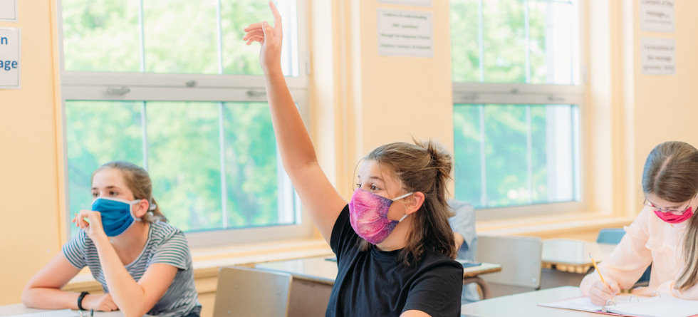 Schülerin in Klassenraum meldet sich und trägt eine Alltagsmaske.
