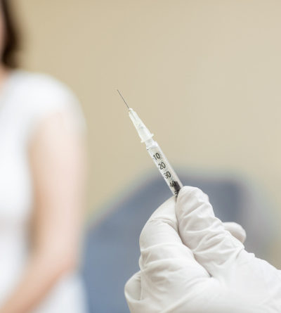 Person hält Impf-Spritze in der Hand. Auf der Behandlungsliege sitzt eine schwangere Frau.