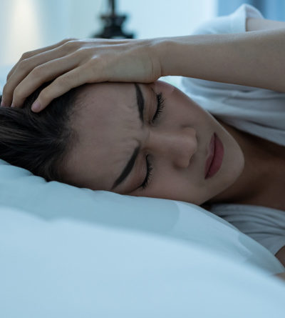 Frau mit schmerzverzerrtem Gesicht liegt in einem Bett und hält sich den Kopf