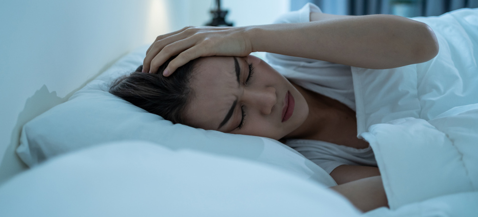 Frau mit schmerzverzerrtem Gesicht liegt in einem Bett und hält sich den Kopf