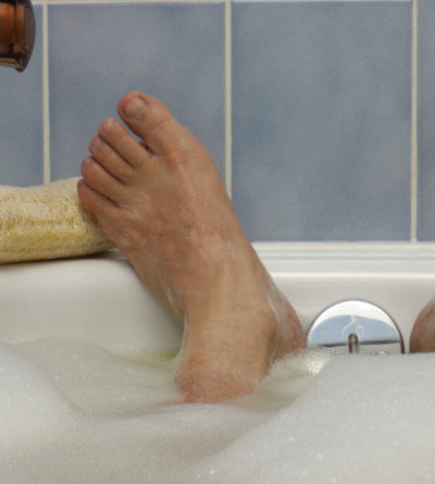 Die Füße eines Mannes, die aus einem Schaumbad ragen, in das seitlich Wasser einläuft.