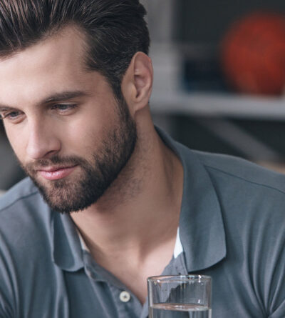 Mann hält ein Glas Wasser und schaut auf eine Tablette, die er in der anderen Hand hält.