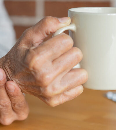 Alte Person hält sich mit der einen Hand am Handgelenk und hält in der anderen Hand eine Tasse.