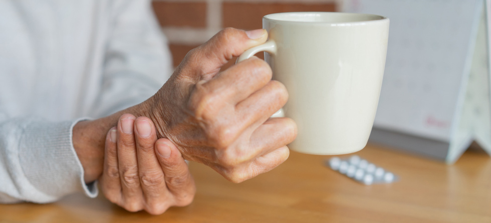 Alte Person hält sich mit der einen Hand am Handgelenk und hält in der anderen Hand eine Tasse.