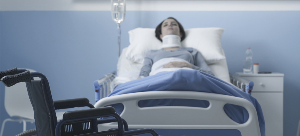 Frau liegt in einem Bett im Krankenhaus. Trägt eine Halskrause. Vor dem Bett steht ein Rollstuhl.