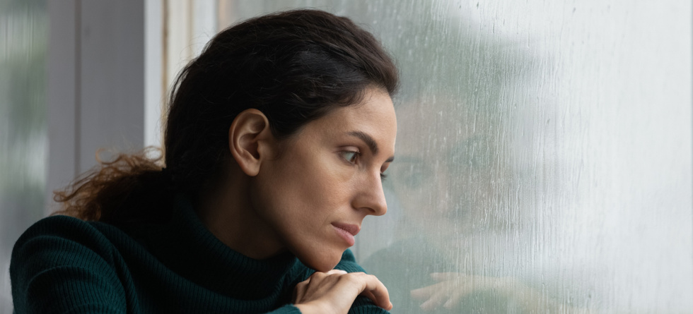 Frau sitzt vor einem Fenster und sieht traurig an die Scheibe, an der Regentropfen herunterlaufen.