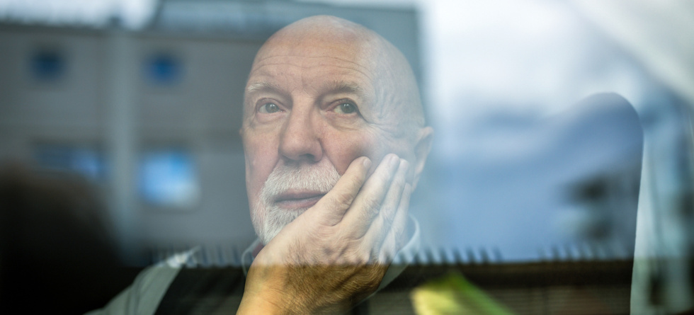 Portrait eines nachdenklichen Senioren, der sich mit der Hand an sein Kinn fasst und durch ein Fenster schaut.