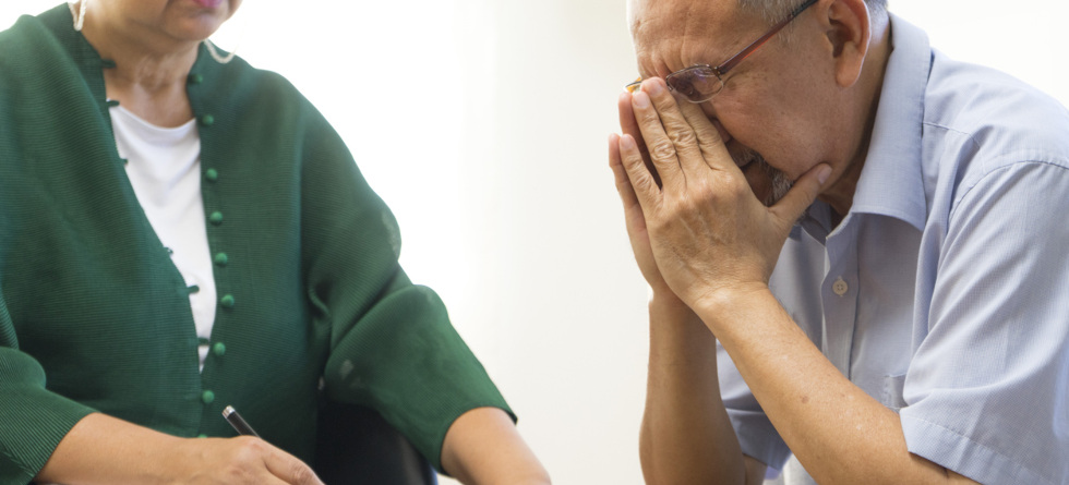 Depressiver älterer Mann teilt seine Probleme während einer Therapiesitzung mit einer Person, die auf einem Klemmbrett schreibt.