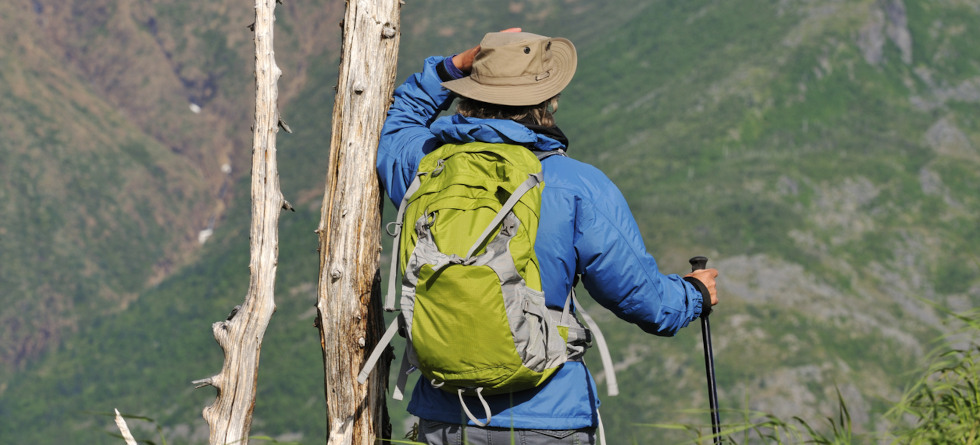Person mit Outdoor-Jacke und Rucksack steht auf einem Berg und blickt ins Tal.